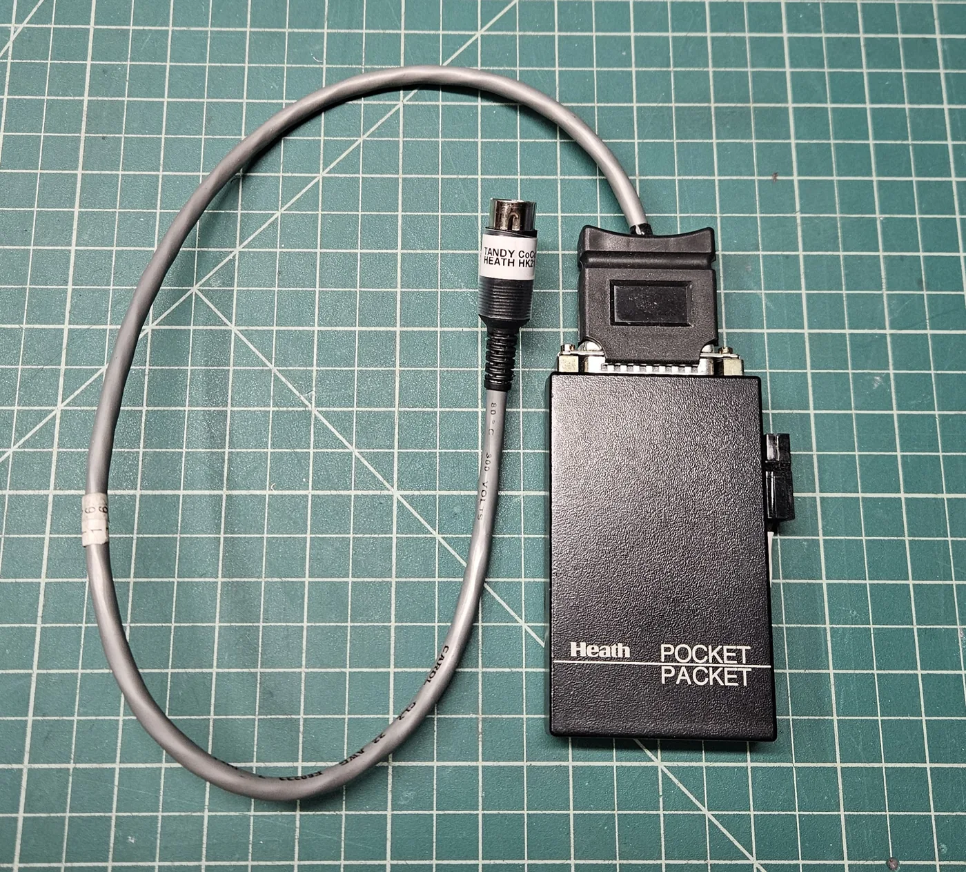 The Heath Pocket Packet with a gray cable plugged into its DB-25 connector, ending in a round 4-pin DIN plug with a label