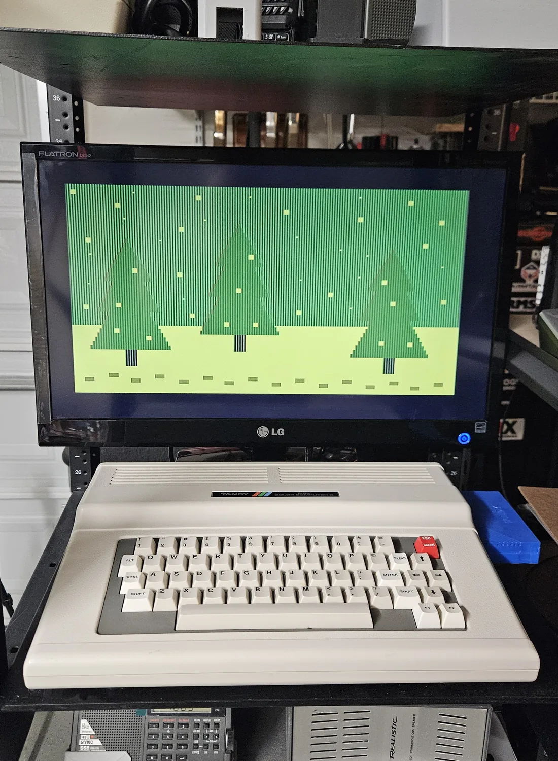 The monitor above the Color Computer 3 showing a graphics demo of green pine trees in falling snow
