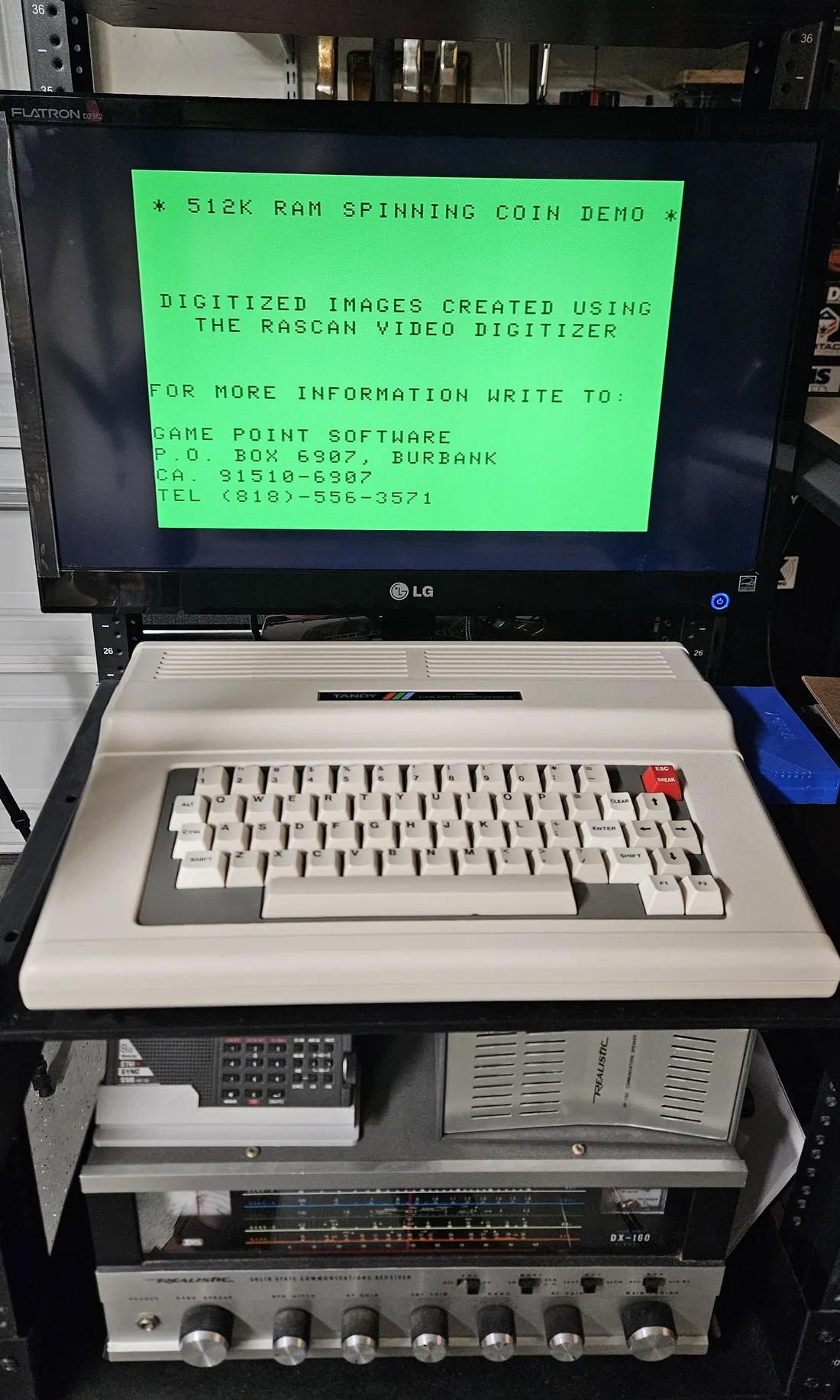 The Color Computer 3 on a rack shelf under the LG monitor, which shows the title screen of a 512K RAM spinning coin demo; a Realistic DX-160 receiver sits on the shelf below