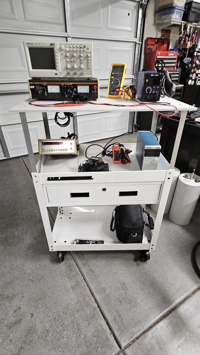 The finished cart standing in the garage with test equipment on the top shelf and the original tray, and a carrying bag on the bottom shelf
