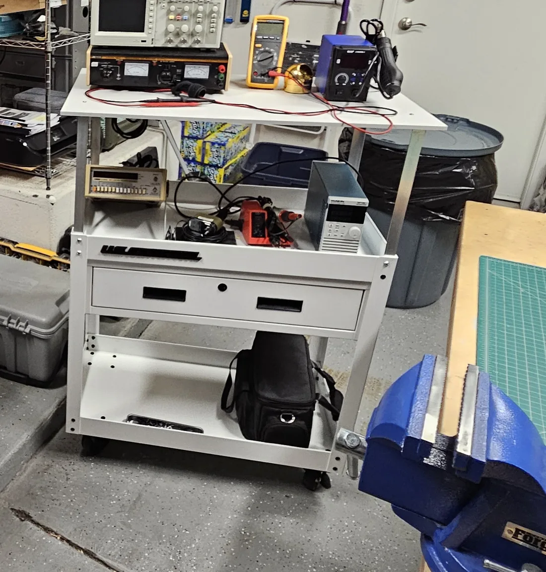 The cart in place in the shop next to a workbench and a blue vise, loaded with an oscilloscope, a power supply, a multimeter, a rework station, a frequency counter and an electronic load