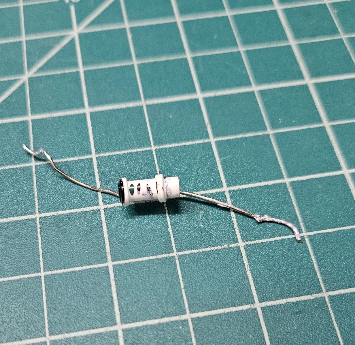 The small white cylindrical part lying on a green cutting mat after it was removed, with a wire lead at each end