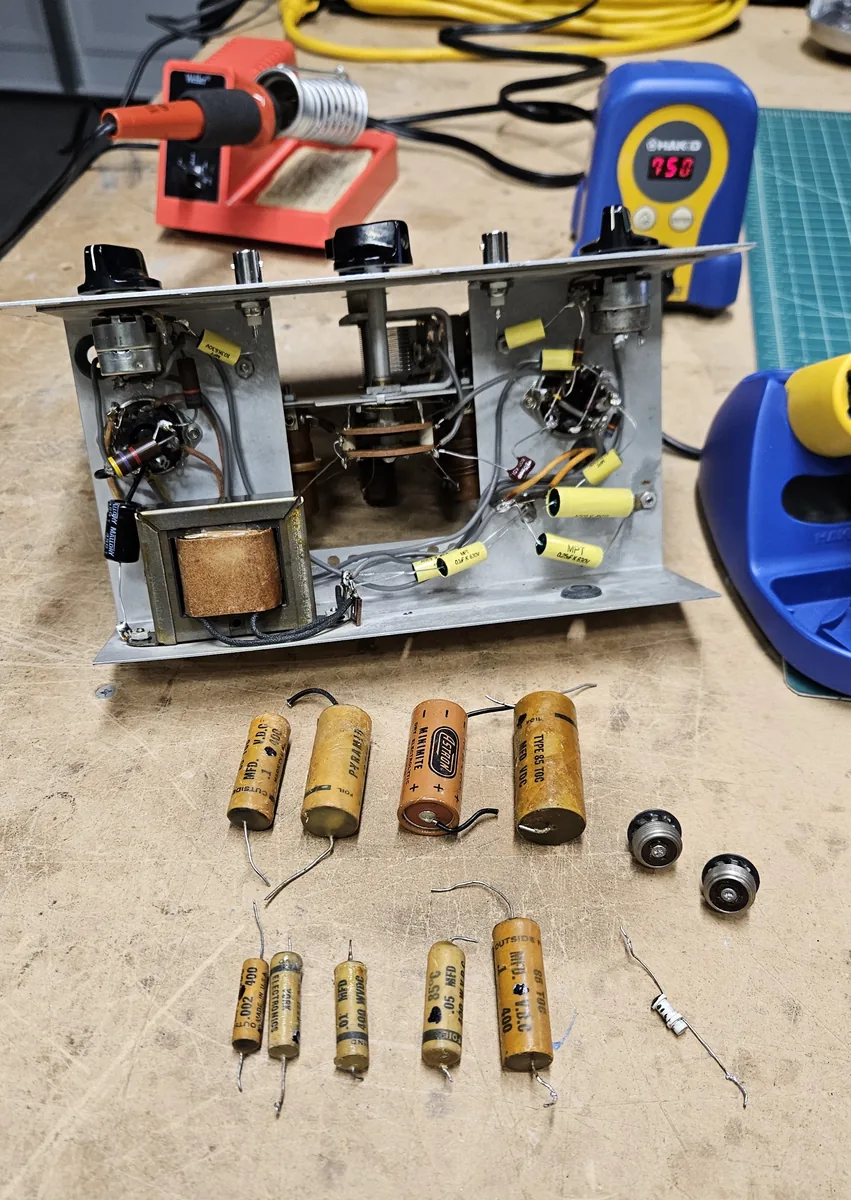 The chassis with new yellow capacitors installed, and in front of it nine old capacitors, two old round connectors and a small white part laid out on the bench, with soldering irons behind