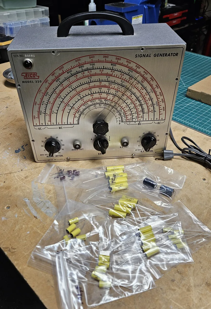 The signal generator on the bench with small plastic bags of new yellow film capacitors, two black electrolytic capacitors and small red mica capacitors laid out in front of it