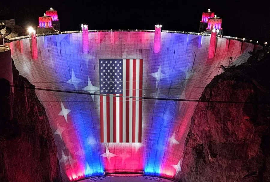 Hoover Dam at night, its face floodlit in red, white and blue with stars, and a large American flag hanging in the middle