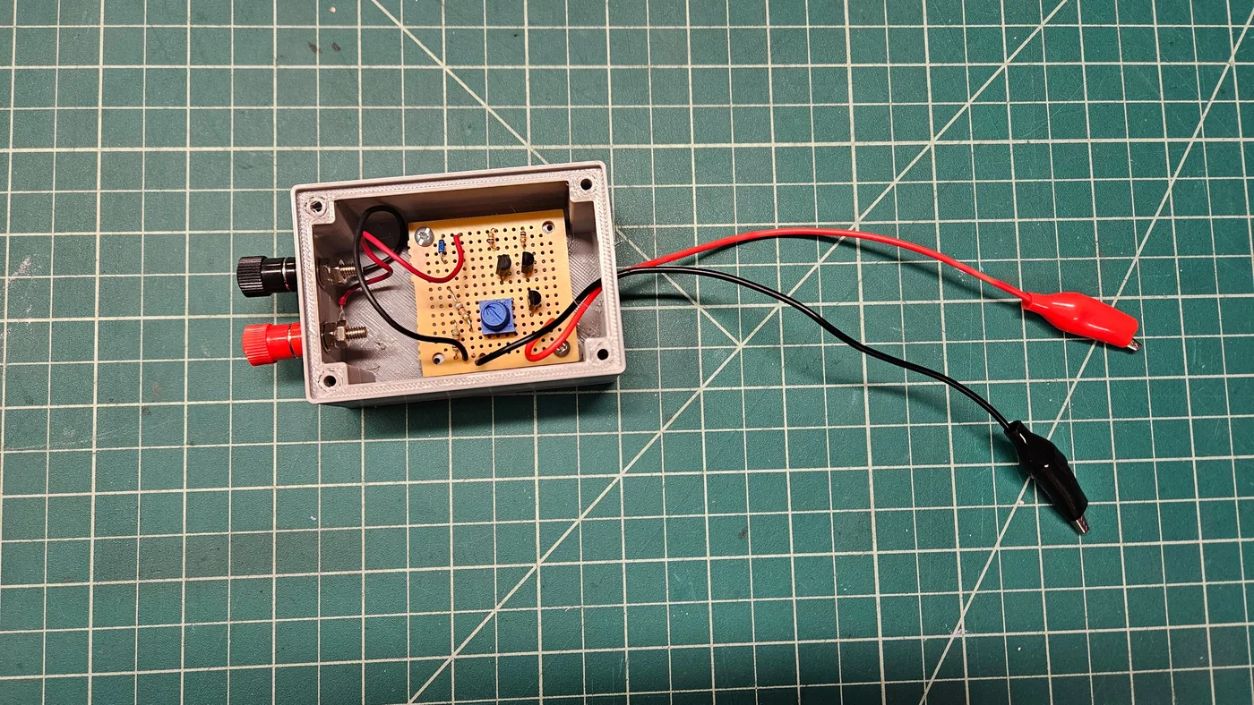 The open gray box with the perfboard inside, red and black terminals on the side, and red and black leads with alligator clips coming out