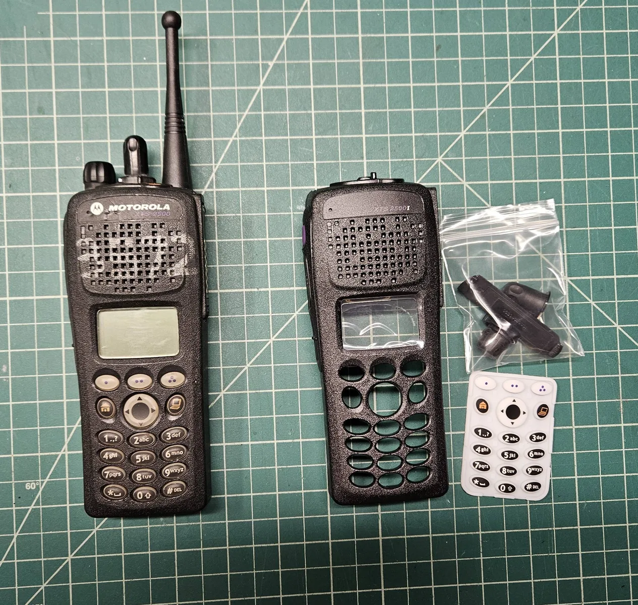 A worn Motorola XTS 2500 with a faded keypad on the left, beside a new black housing, a new white keypad and a bag of new knobs on a cutting mat